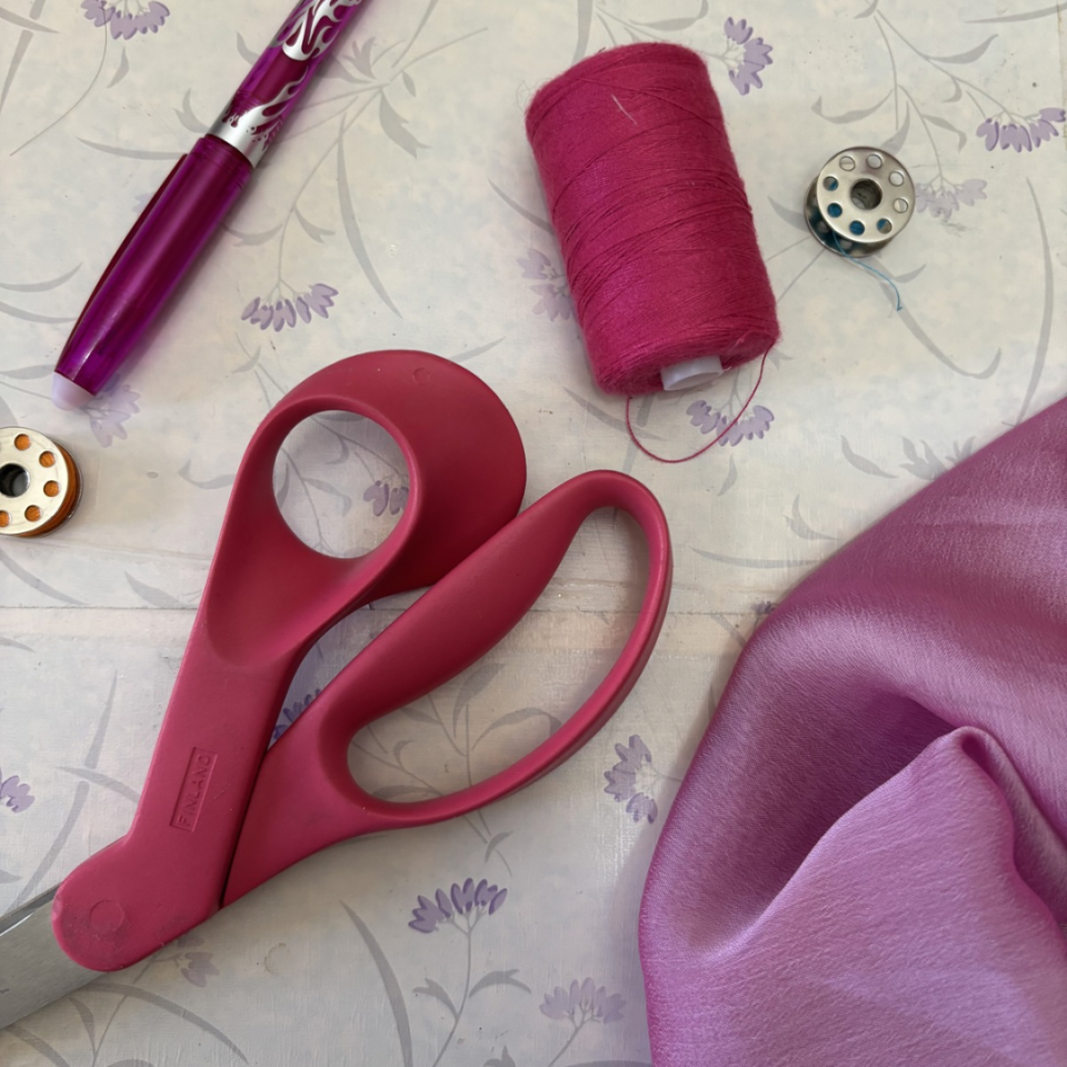 Forbici rosa con vicino tessuto rosa e filo rosa appoggiati al tavolo